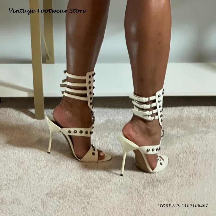 White open toe stiletto heels with silver rivet details and ankle buckle straps for evening wear.