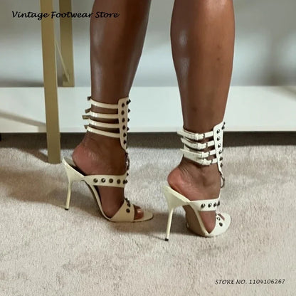 White open toe stiletto heels with silver rivet details and ankle buckle straps for evening wear.