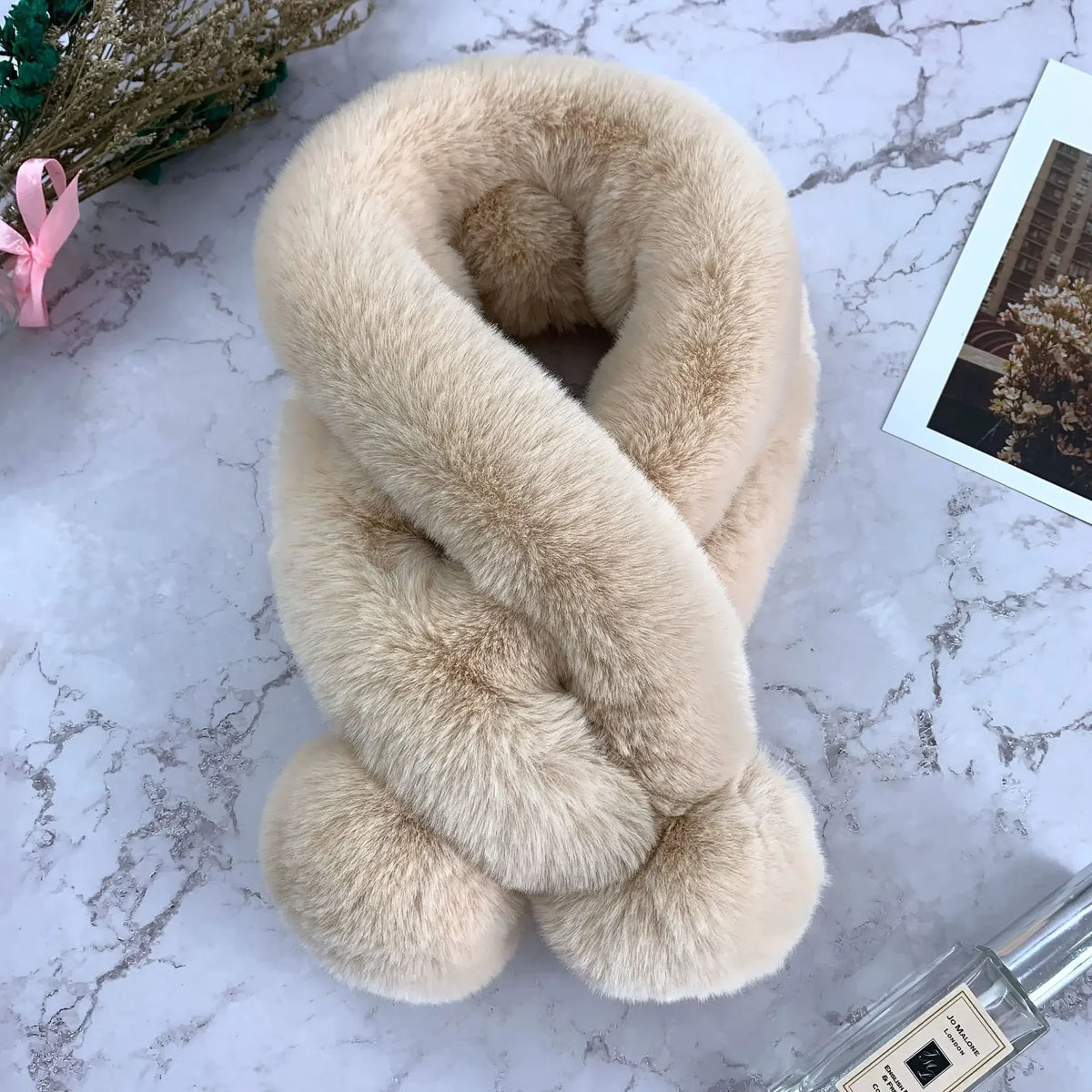Beige plush faux-fur cross scarf with fuzzy ball ends, resting on a marble surface.