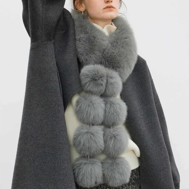 A long grey faux fox fur scarf with plush pom poms worn around the neck.