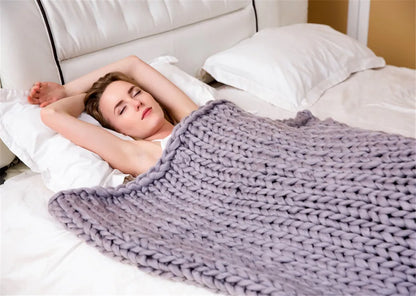 A grey chunky knit blanket made from thick yarn, shown on a bed with a sleeping person.