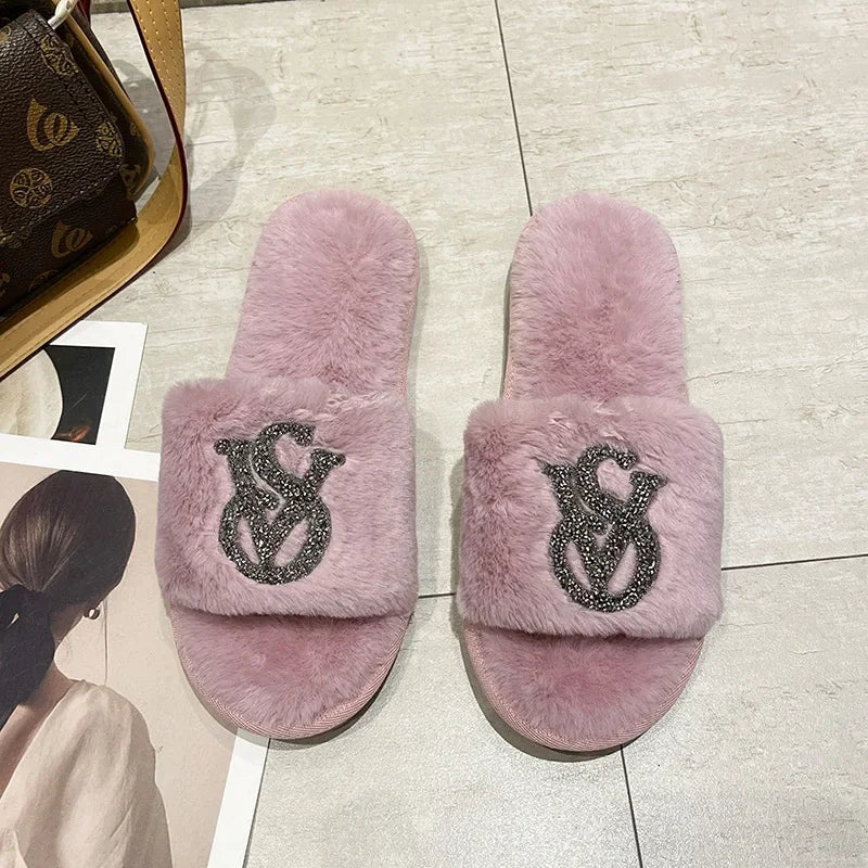 Light pink fuzzy slip-on shoes with rhinestone logos and a non-slip sole for comfortable home lounging.