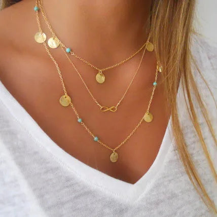 Gold-color multilayer necklace with coin charms, turquoise beads, and an infinity symbol pendant.