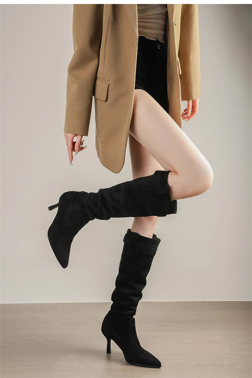 Black suede pointed-toe knee-high boots with thin high heels and buckled strap details.
