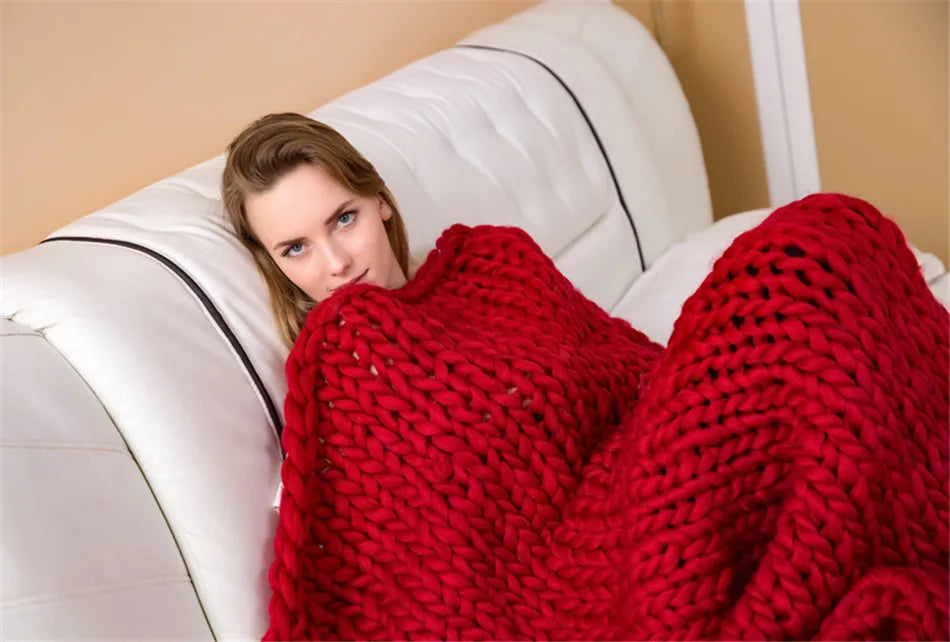 A woman wrapped in a chunky, handmade red yarn knit blanket, lounging on a white sofa.