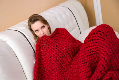 A woman wrapped in a chunky, handmade red yarn knit blanket, lounging on a white sofa.