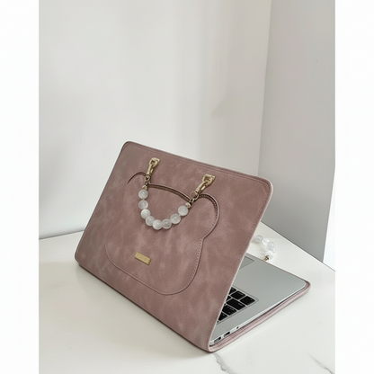 Dusty pink faux-suede laptop case with bead handle and gold hardware on white desk.