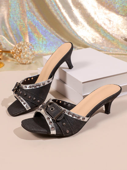Black slide sandals with silver buckle, studs, and kitten heels on a display stand.