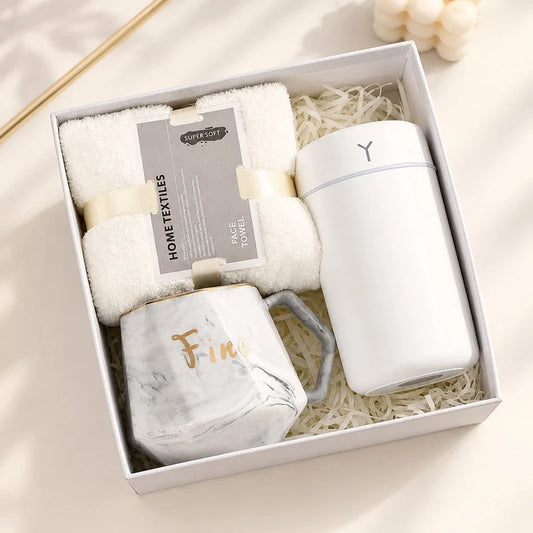 White gift box containing a folded face towel, a marble-patterned mug, and a white tumbler nestled in paper shred.