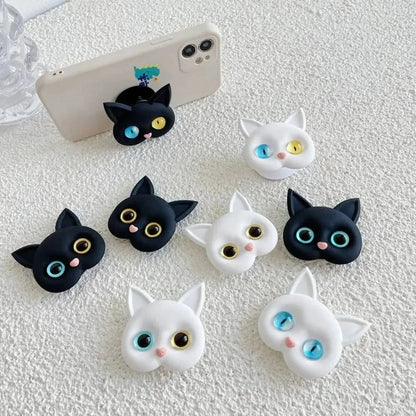 Several 3D cat head phone grips in black and white, with one attached to the back of a light-colored phone.