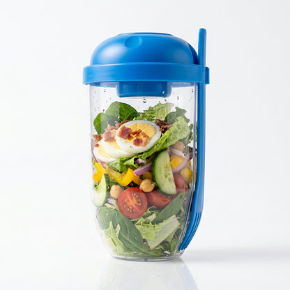Clear salad cup with blue lid and fork, filled with greens, sliced eggs, tomatoes, and cucumbers