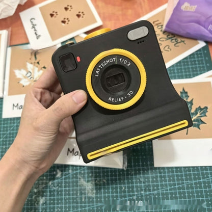 Hand holding a black coffee latte art printing camera with yellow lens and trim, surrounded by design templates.