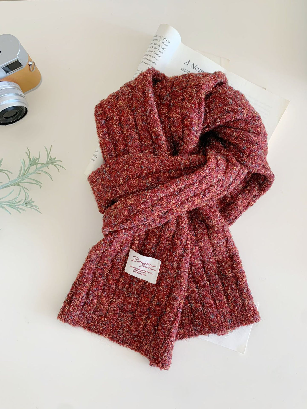 Red knit scarf with a small white label, styled with a camera and book.
