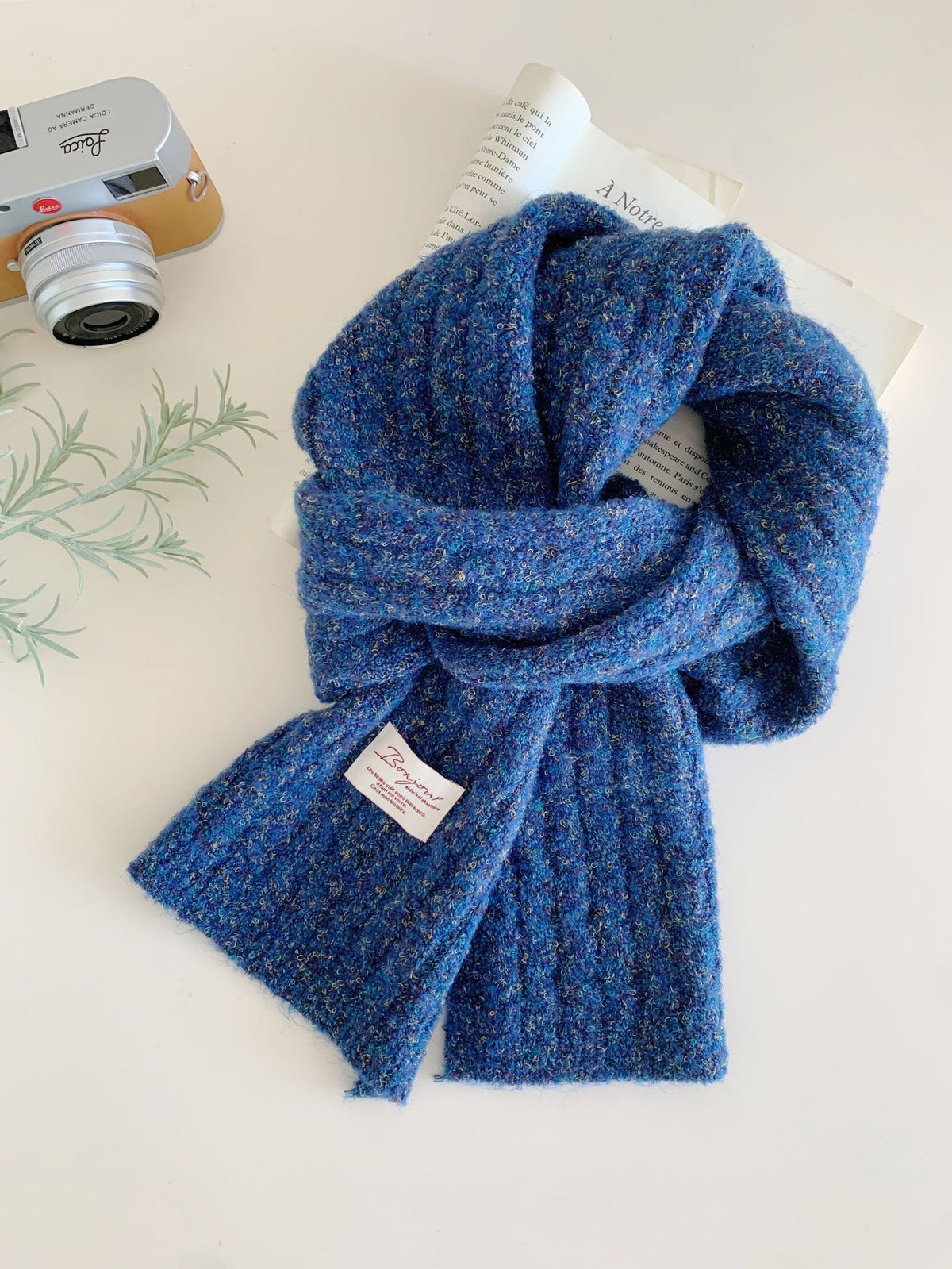 Blue textured knit scarf with a ribbed pattern and a small white tag, styled with a camera and book.
