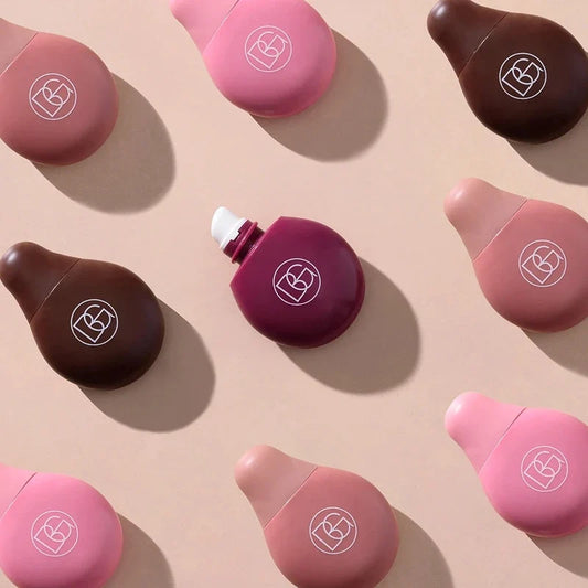 Various lip gloss bottles in shades of pink, brown, and dark purple arranged on a beige surface with one open applicator.