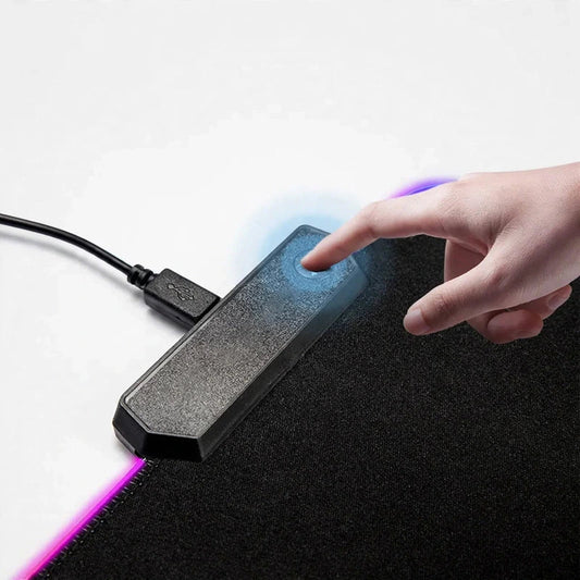 A hand pressing the power button on the control module of an illuminated gaming desk mat connected via USB.