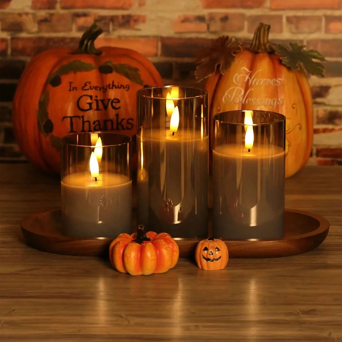 Flameless LED pillar candle set in glass holders with decorative pumpkins on a wooden tray.