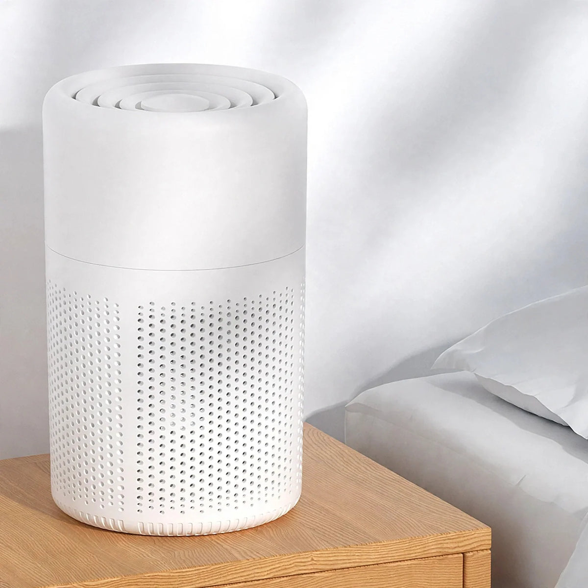 White cylindrical desktop air purifier with perforated intake grille sitting on a wooden nightstand.