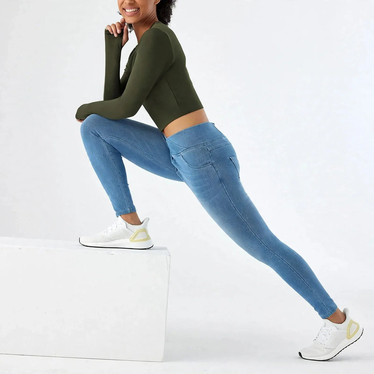 Woman wearing cross-waist skinny blue denim yoga jeans and olive long-sleeve top stretching on a white block.