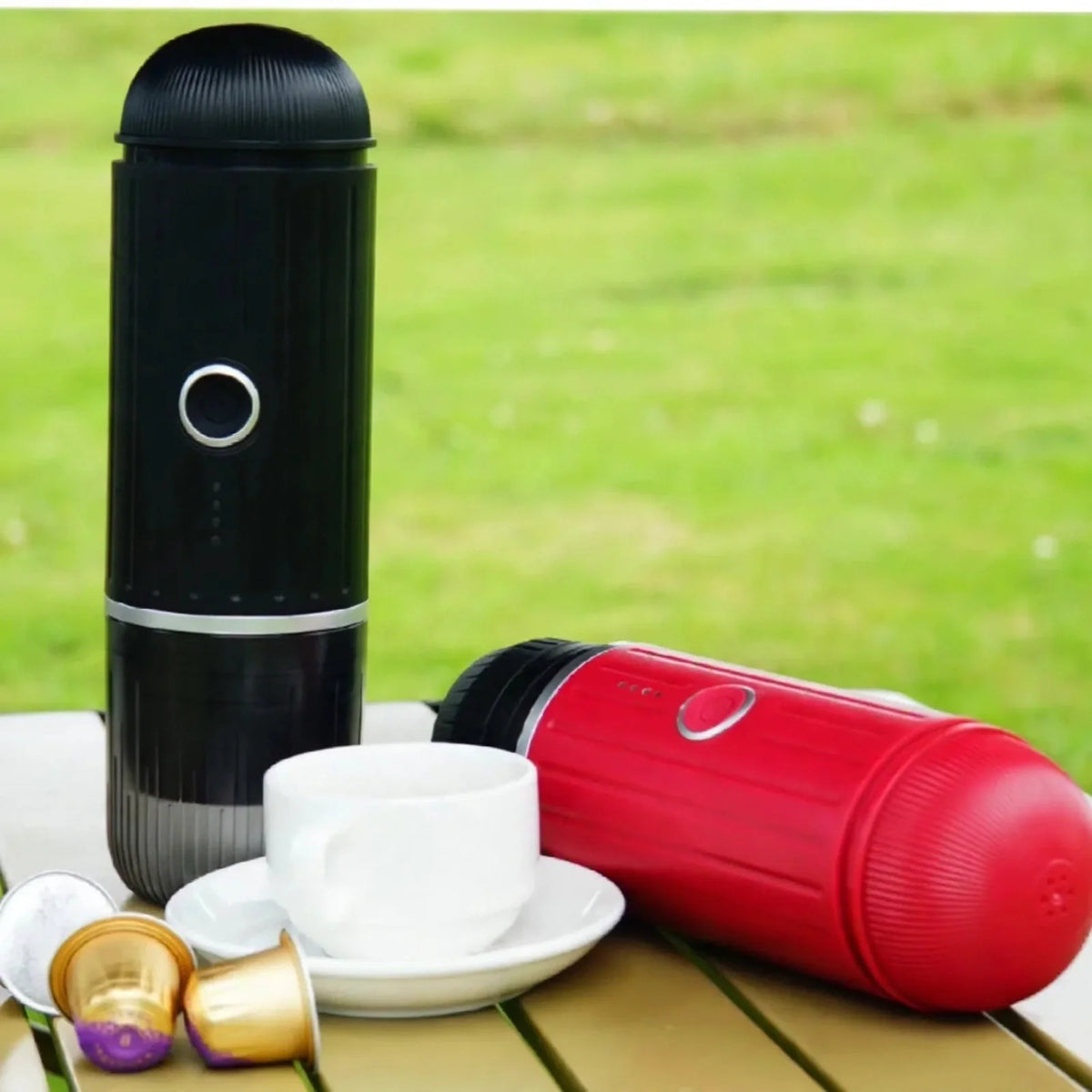 Black and red portable espresso makers on an outdoor table with a white cup and coffee capsules.