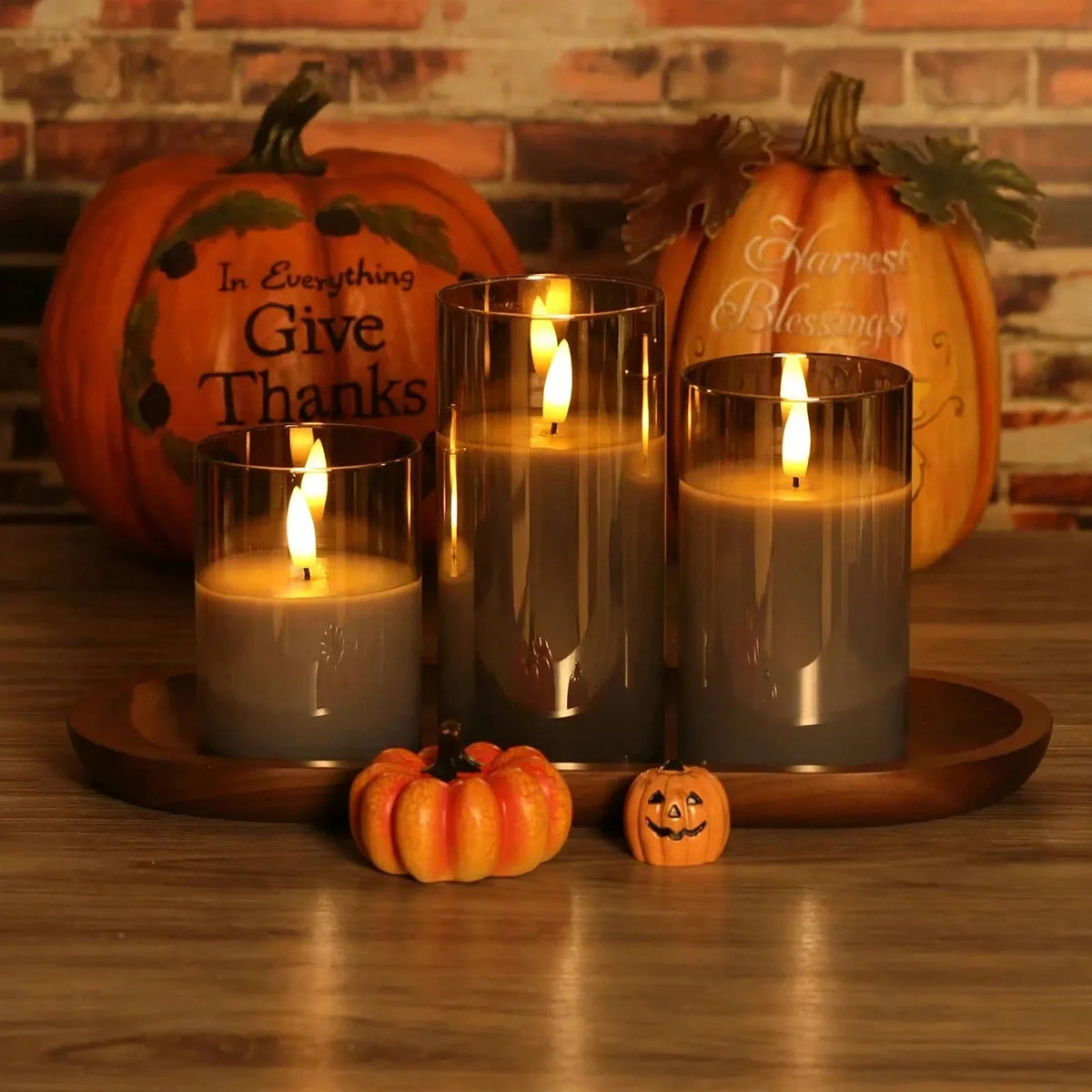 Flameless LED pillar candle set in glass holders with decorative pumpkins on a wooden tray.