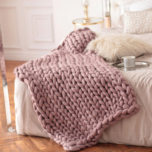 Dusty pink chunky knit throw blanket draped across a bed with decorative pillows and a tray.