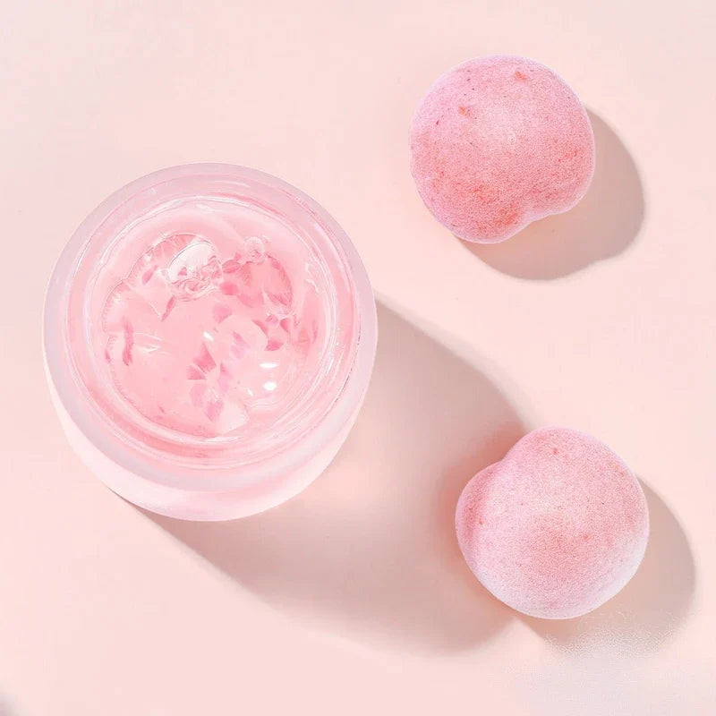 Open jar of pink facial mask with two pink fruit pieces nearby on a solid pink background.
