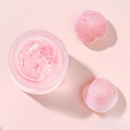 Open jar of pink facial mask with two pink fruit pieces nearby on a solid pink background.