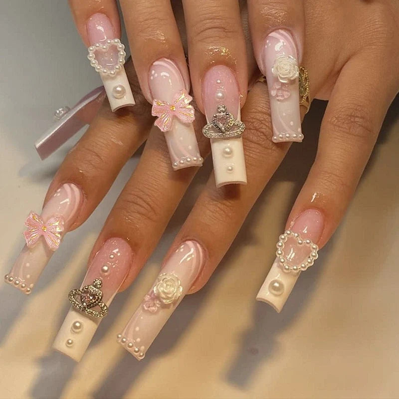 Long square press-on nails with pink and white marbled design, accented with pearls, bows, crowns, and flower charms.