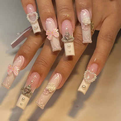 Long square press-on nails with pink and white marbled design, accented with pearls, bows, crowns, and flower charms.