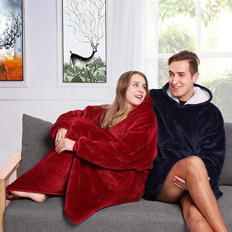 Man and woman sitting on a gray sofa wearing oversized hooded wearable blankets in red and navy blue.