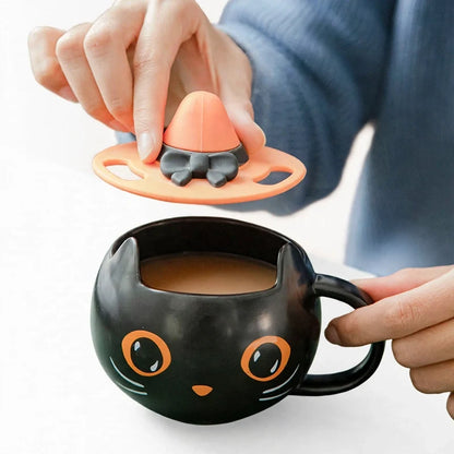 A person placing an orange witch hat lid with a black bow onto a black cat-shaped ceramic mug.