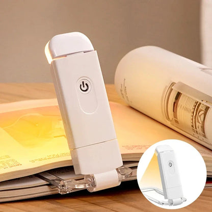 White clip-on book light attached to an open book on a wooden table, with warm light glowing.
