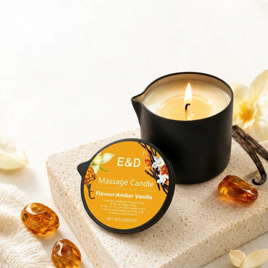 Lit amber vanilla massage candle in a black pour-spout container with amber stones and vanilla flowers.