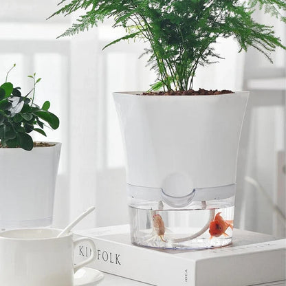 White self-watering planter holding a green plant, with a clear water reservoir below containing small fish.