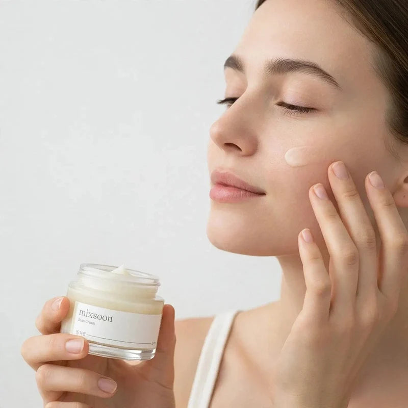 Woman applying white moisturizing cream to her cheek from a glass jar.