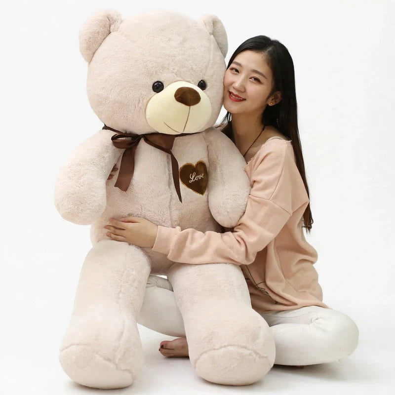 A person hugging a giant beige plush teddy bear wearing a brown ribbon bow and heart patch.