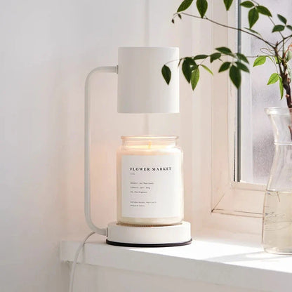 White candle warmer lamp with a jar candle on a windowsill next to a vase with greenery.