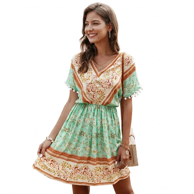 Woman wearing a short-sleeve green and beige floral print mini dress with pom-pom trim and a v-neckline.