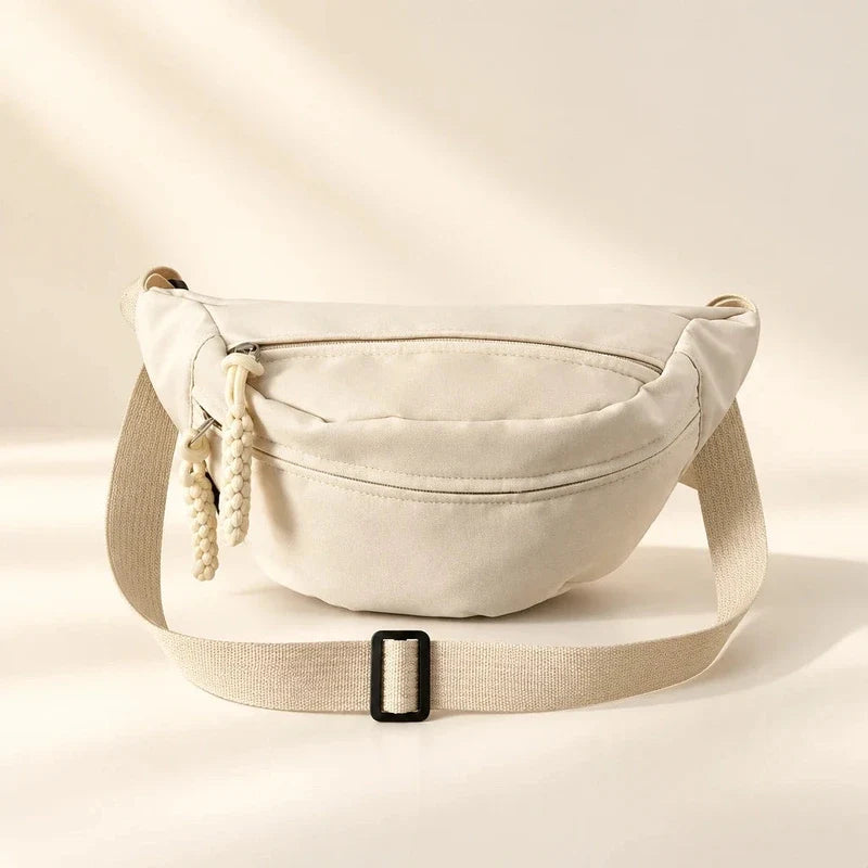 Beige canvas sling bag with front zipper pocket, braided zipper pulls, and adjustable strap with black buckle.
