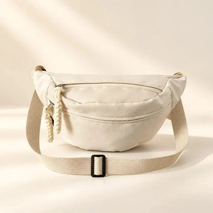 Beige canvas sling bag with front zipper pocket, braided zipper pulls, and adjustable strap with black buckle.