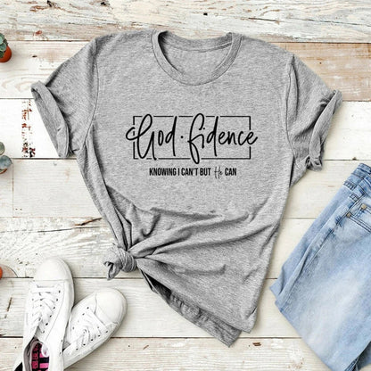 Grey t-shirt with Godfidence text graphic, laid flat on wood with jeans and sneakers.