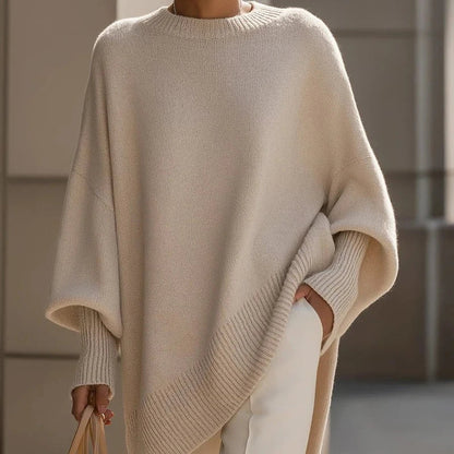 Woman wearing an oversized beige mock neck sweater with batwing sleeves and ribbed cuffs, paired with white pants.