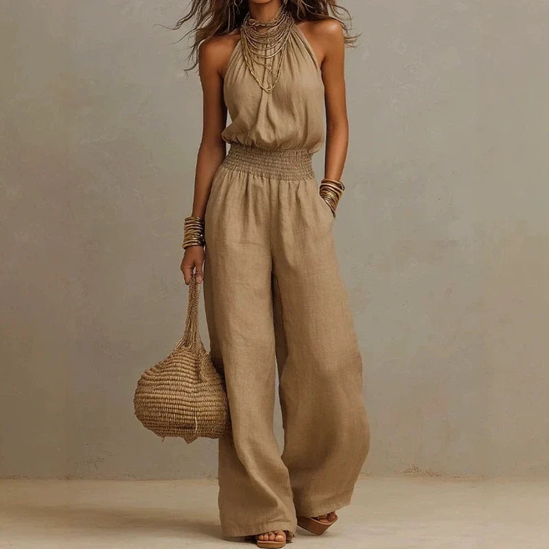 Tan halter wide-leg jumpsuit with a smocked waist, worn with layered necklaces and a woven straw bag.