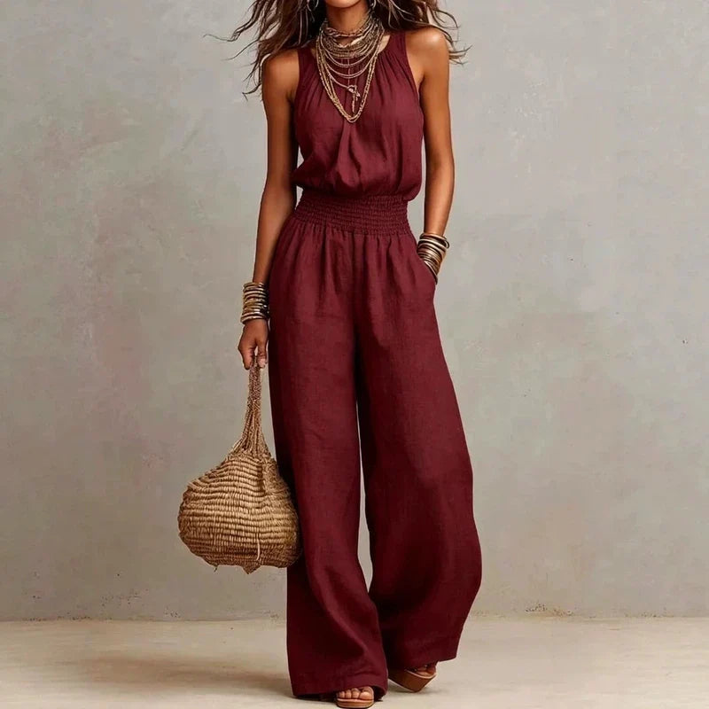 Woman wearing a maroon halter wide-leg jumpsuit with a smocked waist, holding a woven bag.